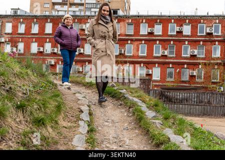 Due donne caucasiche che camminano lungo il percorso attraverso il complesso di alloggi in mattoni rossi, una donna con il cappotto di fossa beige conduce mentre un amico in giacca viola segue, Foto Stock