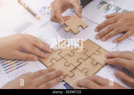 Vista dall'alto di un team aziendale diversificato che collabora per collegare pezzi di puzzle in legno a report finanziari e grafici su una scrivania da ufficio. Busin Foto Stock