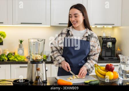 La donna mette la carota grattugiata nel frullatore e affetta la banana sul tagliere per preparare il frullato. Sana colazione con vitamine, ingredienti freschi, routine di cucina casalinga e buona energia. Foto Stock