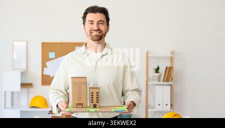Architetto sorridente che tiene in mano il modello di costruzione in cartone in un ufficio moderno, ritratto di un ingegnere o designer maschile che presenta uno sviluppo urbano sostenibile, r Foto Stock