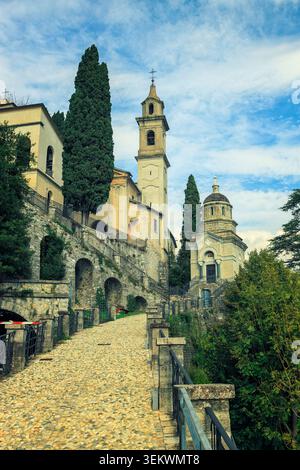 Brienno, Italia - 15.10.2022: Antico complesso religioso caratterizzato da un alto campanile e da una cupola adagiati su un cielo luminoso con vegetazione Foto Stock