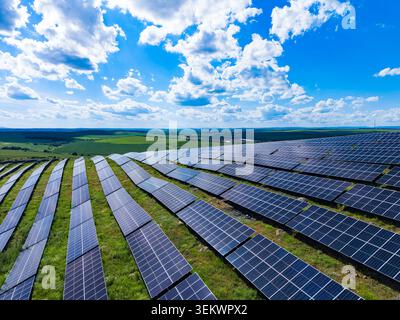 File di pannelli solari sono installati su una collina. I pannelli riflettono la luce solare. I campi verdi si estendono in lontananza sotto un cielo blu con nuvole. IT Foto Stock
