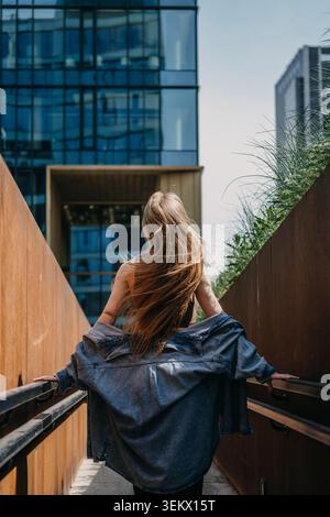 Una donna cammina su una rampa pedonale tra le pareti guardando verso un moderno grattacielo di vetro. Infrastruttura urbana percorribile a piedi, percorso pedonale urbano, intelligente Foto Stock