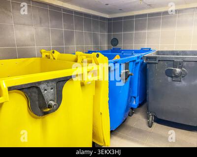 Area interna per la raccolta dei rifiuti con grandi contenitori per rifiuti in plastica nei colori giallo, blu e nero per una separazione sostenibile dei rifiuti Foto Stock