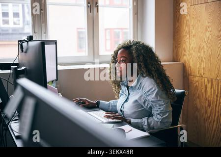 Esperta di tecnologia donna dai capelli ricci che lavora su un doppio computer in una startup Foto Stock