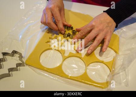 Le mani della persona tagliano fiocchi di neve e forme circolari dall'impasto crudo per biscotti con i taglierini Foto Stock