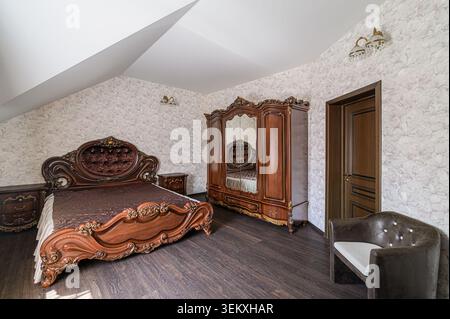 lussuosa camera da letto in un ambiente attico presenta eleganti mobili di legno scuro, carta da parati a motivi geometrici e una finestra con tende marroni. La camera è bagnata Foto Stock