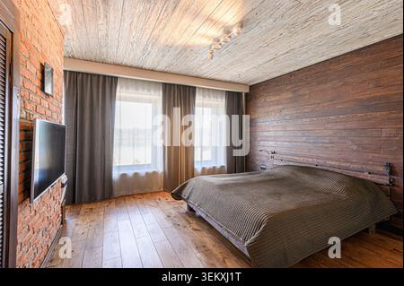 Accogliente camera da letto con pareti in mattoni, accenti in legno, TV moderna e luce naturale che filtra attraverso tende trasparenti. La camera ha un'atmosfera calda e invitante Foto Stock