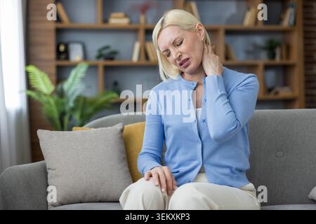 Una donna bionda matura seduta su un divano, che soffoca e si strofina il collo doloroso con una mano, mostrando rigidità e dolori muscolari dovuti a dolori cronici al collo o postura inadeguata a casa Foto Stock