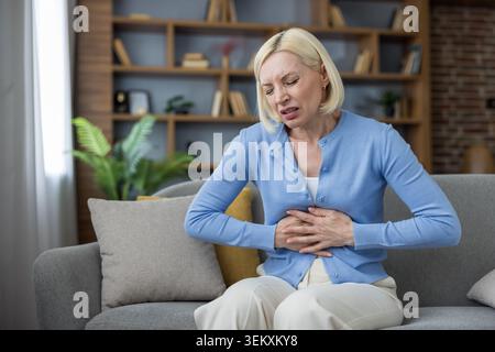 Donna anziana che soffre di grave dolore addominale e indigestione, che tiene le mani sullo stomaco mentre siede su un comodo divano in un appartamento moderno, si sente male e male Foto Stock