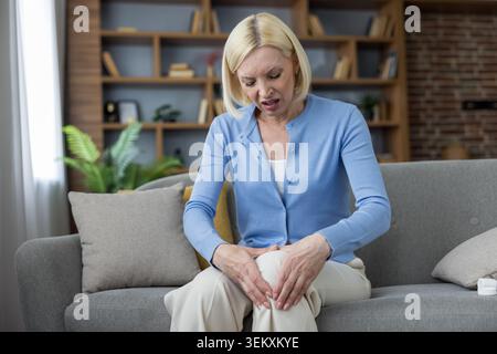 Donna anziana seduta su un divano, che soffre di dolori improvvisi al ginocchio, che tiene in mano le articolazioni dolorose, illustrando problemi di salute legati all'età e disagio nella vita quotidiana Foto Stock