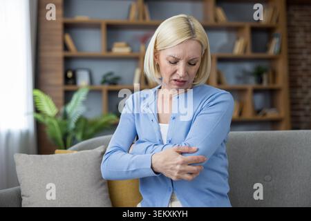 Donna matura che soffre di disagio con il gomito, sfregando delicatamente l'articolazione con la mano mentre si siede su un divano in un ambiente accogliente, indicando dolore o lesioni Foto Stock