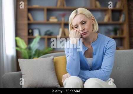 Donna anziana che soffre di malinconia, solitudine e tristezza, soffre di stress emotivo e problemi di salute mentale mentre si siede su un divano nel suo salotto Foto Stock