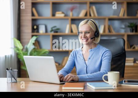 Donna matura sorridente che indossa le cuffie con microfono impegnata attivamente nel lavoro da remoto, partecipando a una riunione online o fornendo assistenza clienti dal suo ufficio domestico Foto Stock