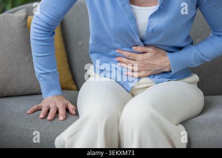 Donna irriconoscibile che tiene la mano sullo stomaco sentendo crampi o problemi digestivi, seduto su un divano a casa, soffre di mal di stomaco, dolore del periodo o disagio mestruale Foto Stock