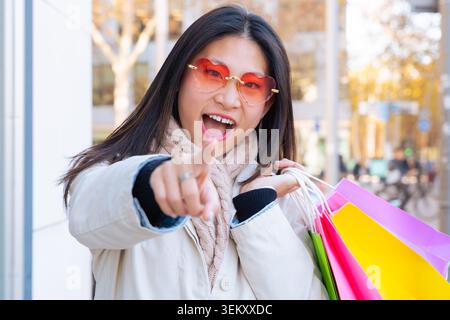 Donna asiatica con occhiali da cuore che puntano alla macchina fotografica, tenendo in mano borse colorate Foto Stock