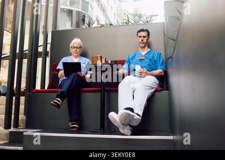 Vista ad angolo basso degli operatori sanitari maschili e femminili che utilizzano tecnologie wireless durante la pausa caffè in un moderno ospedale Foto Stock