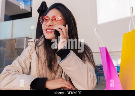 Felice donna asiatica che parla su smartphone con borse dello shopping in un'area caffetteria all'aperto Foto Stock
