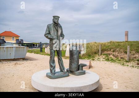 Scultura in bronzo del marinaio solitario con il suo borsone a Utah Beach, in Normandia. Lo sfondo presenta dune di sabbia, l'obelisco commemorativo, una casa con wi Foto Stock