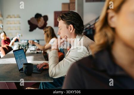 Uomo d'affari con la mano sul mento che lavora su un computer portatile seduto alla scrivania in un'area di lavoro condivisa Foto Stock