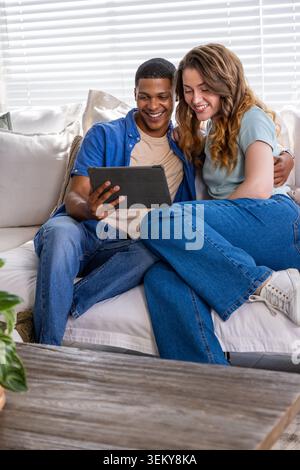 Coppie diverse sedute su un divano leggero nel soggiorno, che condividono il tablet e indossano jeans e t-shirt Foto Stock