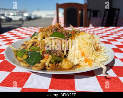 Piatto di tagliatelle udon saltate con manzo, cavolo e cipolle verdi servite con verdure sottaceto. Impostazione esterna su una scheda a scacchi rossa e bianca Foto Stock
