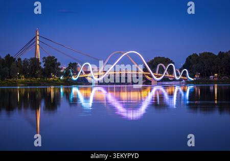 Ponte pedonale Obolon illuminato a Kiev, Ucraina. Le luci che cambiano colore si riflettono sull'acqua calma, creando un suggestivo paesaggio urbano notturno e riflessi Foto Stock
