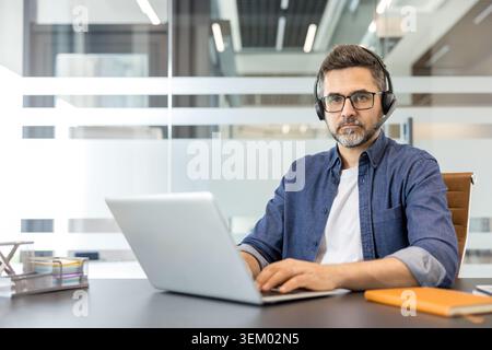 Uomo maturo con cuffie e occhiali che lavora su un laptop alla sua scrivania in un ufficio moderno, fornendo supporto tecnico e servizio clienti con un'espressione mirata Foto Stock