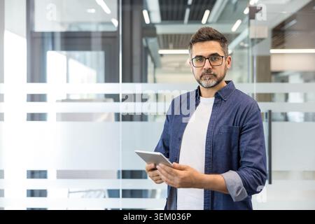 Professionista maschile sicuro di occhiali da vista e camicia in denim che regge un tablet in un ufficio moderno con pareti divisorie in vetro, che rappresenta tecnologia mobile, comunicazione e produttività Foto Stock