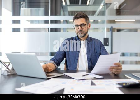 Un uomo d'affari sicuro di sé esamina documenti e tipi su un notebook in una moderna scrivania, concentrandosi su report finanziari, grafici e pianificazione dei progetti per il successo della gestione Foto Stock