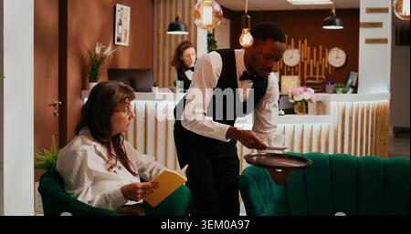Il Bellhop serve una tazza di caffè a una donna che si rilassa nell'area lounge, mentre gli ospiti senior dell'hotel si godono una bevanda rinfrescante e leggono un libro. Dipendente che fornisce servizi all inclusive durante le vacanze. Foto Stock