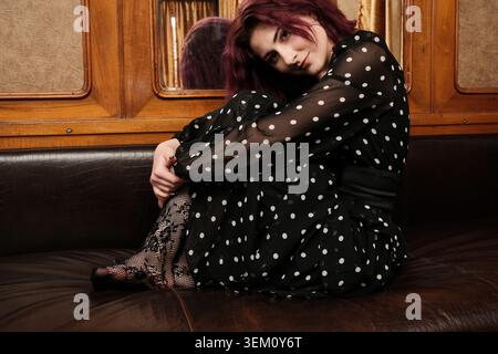 Una donna siede con le gambe arricciate e indossa un elegante abito nero a pois. Foto Stock