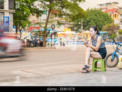 Vietnam - 11 giugno 2025: Vicino al quartiere vecchio e accanto al fiume Rosso, una donna che lavora in un bar di strada usa il suo telefono vicino alla strada, vicino alla città di tra Foto Stock