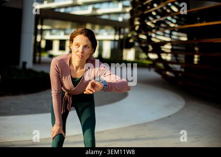 Una donna si piega in avanti, guardando il suo orologio mentre fa jogging in un parco cittadino. Indossa abiti da ginnastica e mostra di concentrarsi sulla sua routine di allenamento. T Foto Stock