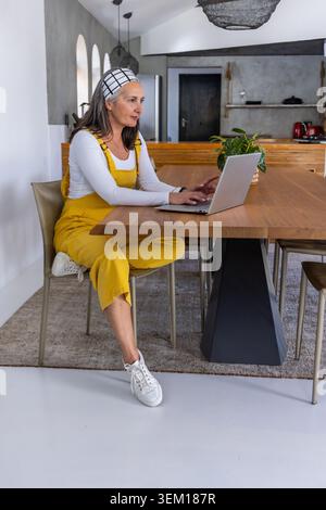 Donna di mezza età in tuta gialla seduta al tavolo da pranzo tenendo in mano la pianta e scrivendo sul laptop Foto Stock