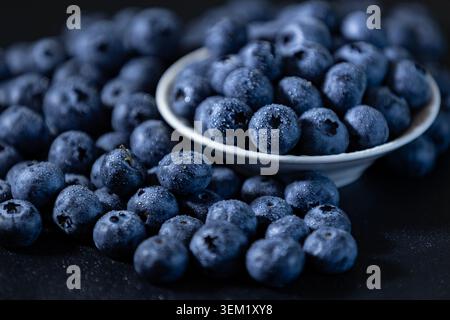 Primo piano di foto macro di mirtilli freschi e succosi ricoperti di gocce d'acqua. Alcune bacche si trovano in una piccola ciotola di ceramica bianca, mentre altre sono sc Foto Stock