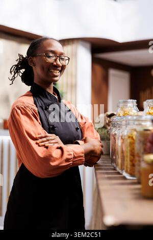 Immagine dettagliata di una donna afroamericana che fa shopping con un grembiule sorridente davanti alla macchina fotografica vicino a vasetti di vetro. Ritratto di una donna nera che indossa occhiali con il braccio incrociato sul petto. Foto Stock