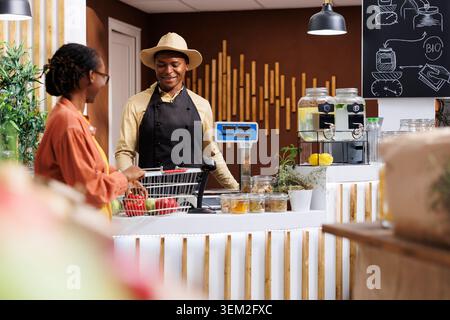 Il cordiale cassiere nero con un sorriso caldo assiste la cliente con i suoi acquisti al banco cassa del mercato locale. L'immagine mostra un eccellente servizio clienti e un'esperienza di acquisto diversificata. Foto Stock