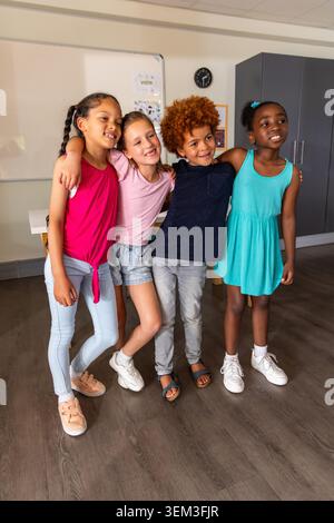 Quattro bambini in piedi con le braccia intorno, in posa in classe vicino alla lavagna bianca con orologio grigio Foto Stock
