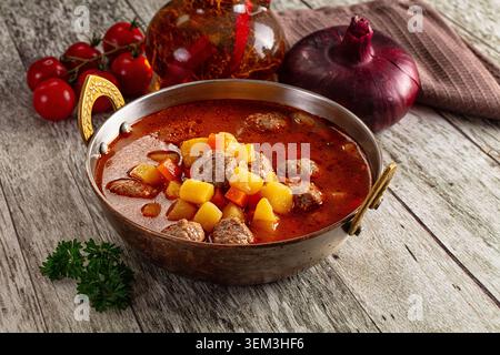Abbondante zuppa di pomodoro con polpette, patate e carote in una ciotola di rame su un tavolo rustico in legno, cipolla rossa e pomodori ciliegini sullo sfondo, primo piano Foto Stock