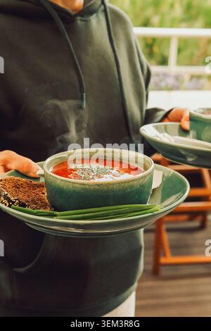 Zuppa calda servita con pane e cipolle verdi in un ambiente informale durante un pomeriggio di sole. la persona tiene due piatti con borsch e pane in re Foto Stock