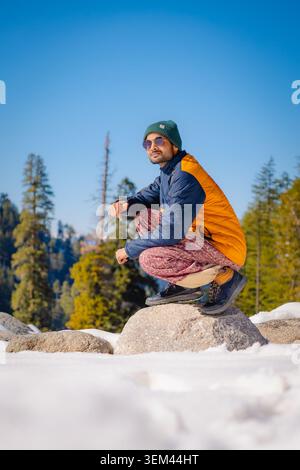 Un giovane uomo che indossa un berretto e una giacca accovacciati su una roccia in un paesaggio innevato di montagna nel Kashmir, India. Foto Stock