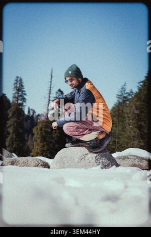 Un giovane uomo che indossa un berretto e una giacca calda accovacciato su una roccia innevata in una pineta, Kashmir, India. Foto Stock