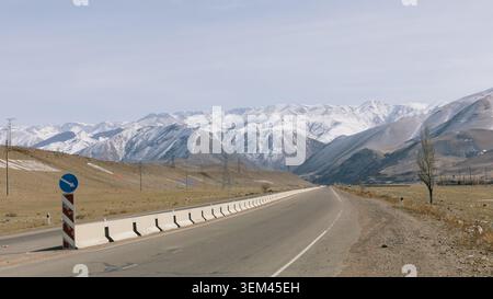 Ampia vista panoramica di una strada asfaltata vuota con barriera di sicurezza che conduce verso le montagne innevate di Tian Shan, arido paesaggio della valle e cielo limpido a Ky Foto Stock