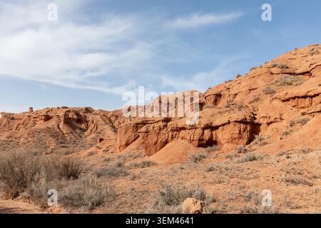 Ampia vista del Mars Canyon con scogliere di arenaria rossa, arbusti secchi e creste erose sotto un cielo blu, Kirghizistan. Destinazione geologica panoramica nella zona centrale A. Foto Stock