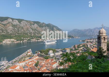 Ampia vista panoramica su Cattaro, la baia di Cattaro e le montagne circostanti in Montenegro, con una grande nave da crociera sull'acqua, tetti rossi e una chiesa Foto Stock