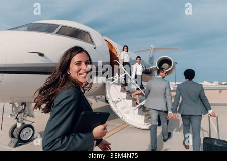 Giovane donna d'affari professionista in un blazer della marina con un tablet digitale saluta i dirigenti aziendali in arrivo con un sorriso caloroso in un'azienda privata bianca Foto Stock