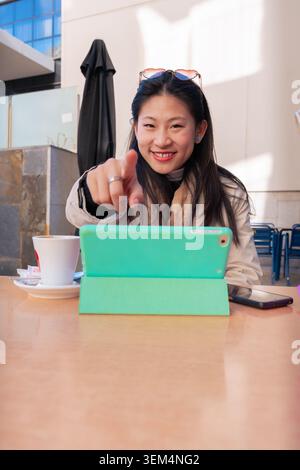 Felice donna asiatica che punta alla fotocamera durante la videochiamata al caffè all'aperto con borse Foto Stock