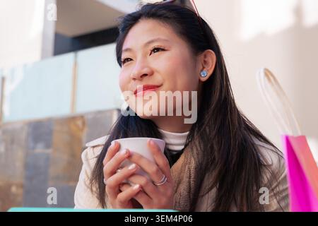 Donna asiatica sorridente che tiene una tazza di caffè all'Outdoor Cafe con borse per lo shopping rosa Foto Stock