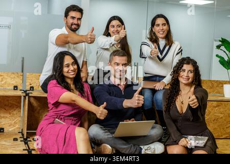 Un gruppo eterogeneo di sei persone celebra il successo di una squadra in un moderno spazio di coworking. Sono riuniti intorno a un notebook, sorridenti e donanti Foto Stock